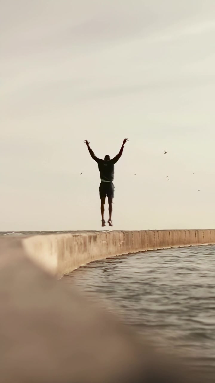 Video verticale: Uomo con top scuro e pantaloncini che inizia con le braccia alzate, salta lungo un molo in riva al mare mantenendo l'equilibrio.