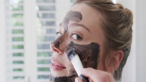 Portrait-of-happy-biracial-woman-applying-beauty-face-mask-in-bathroom,-slow-motion