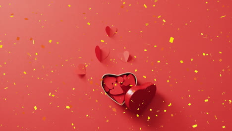 animation of yellow confetti falling over heart paper cutouts in heart shaped box on red background