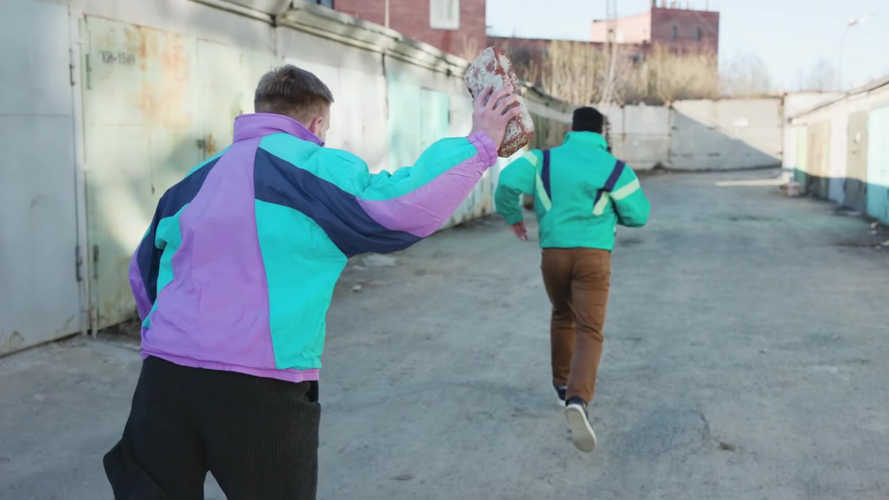 Dos amigos corriendo por un callejón de garajes mientras uno persigue al otro, chaquetas retro coloridas, concreto urbano, lanzado.