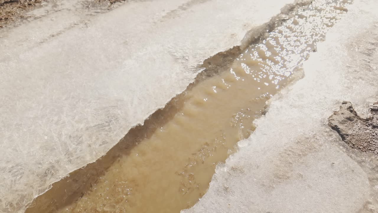 Primo piano di ghiaccio che si scioglie con acqua marrone che scorre, creando una texture astratta naturale. Dettaglio del disgelo invernale, concetto di cambiamento climatico e sfondo di transizione stagionale