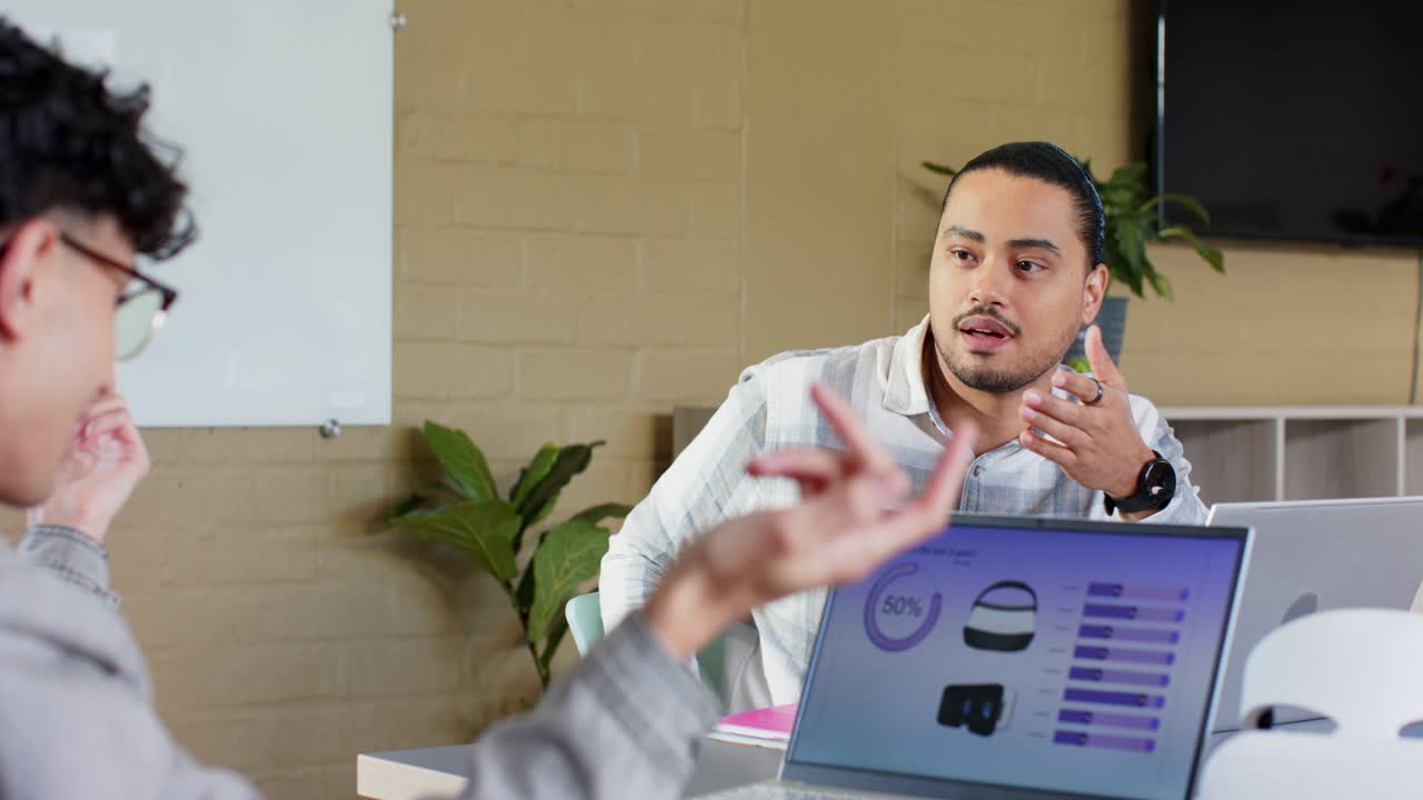 Male colleague listening, responding to gesturing peer pointing laptop charts in office, copy space