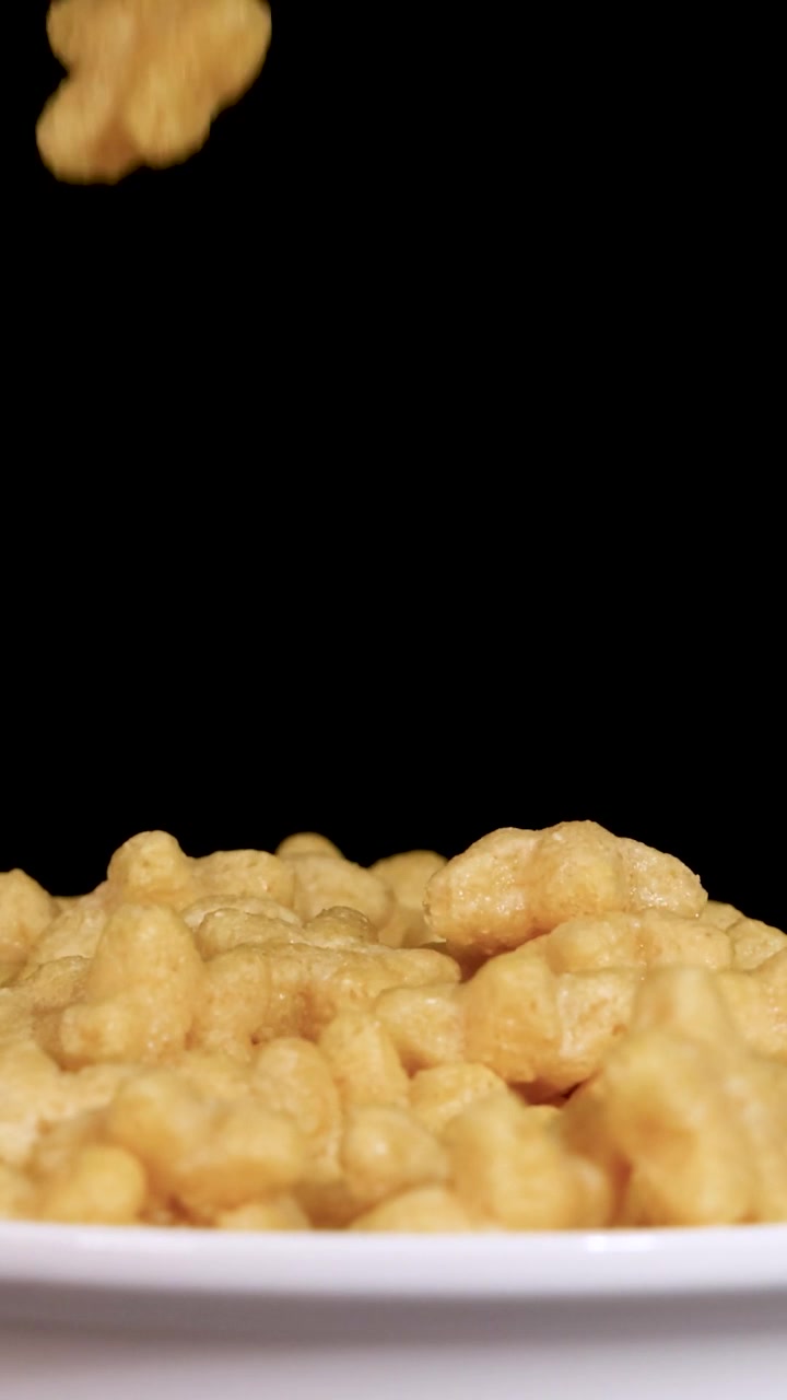 Pezzi di cereali a forma di stella dorati cadono in una ciotola bianca su uno sfondo nero uniforme.