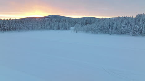 Norbotten-Swedish-Lapland-Polar-circle-aerial-view-glowing-golden-sunrise-over-snow-covered-woodland-and-ice-lake
