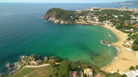 aerial video revealing la fosca beach's pristine turquoise waters, golden sands, and en plaja castle nestled along palamos coastline in spain's costa brava region