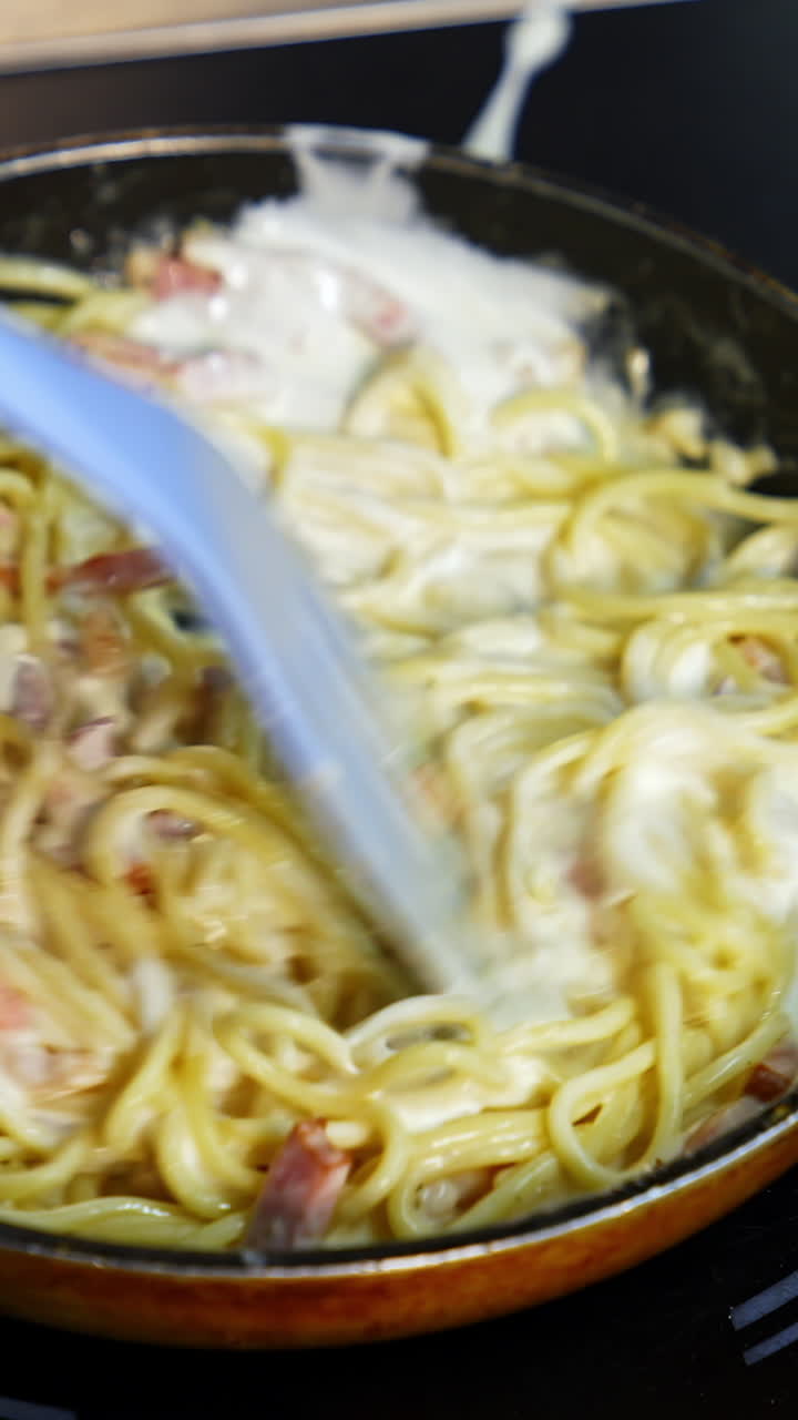 Preparation of a delicious dish in frying pan. Cooking and stirring the spaghetti with white sauce in the frying pan. Close-up. Vertical video