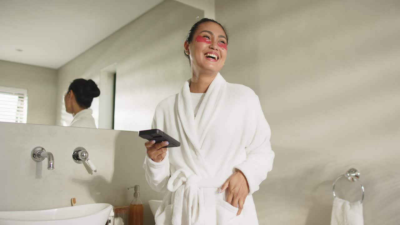 Asian woman reacting to message, raising smartphone and laughing, wearing eye patches beside sink