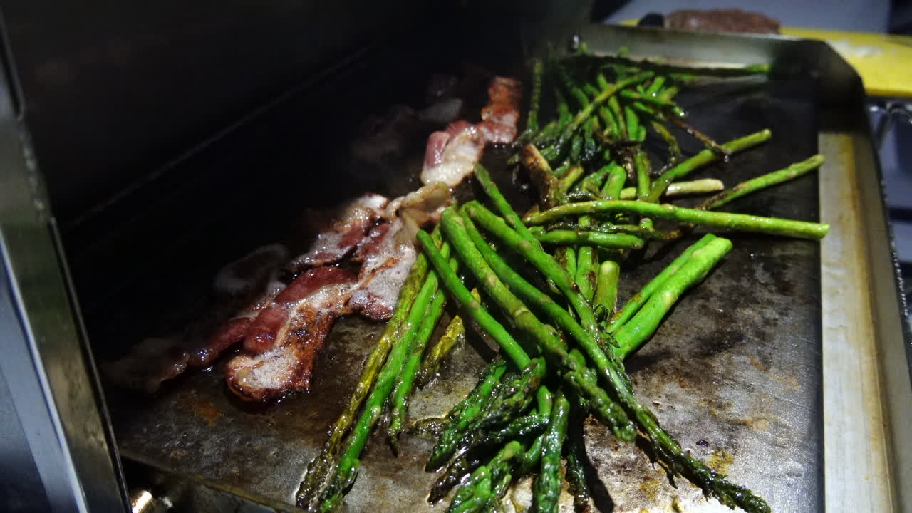 Steady shot of a grill opened on a sail boat, cooking asparagus and bacon on a griddle