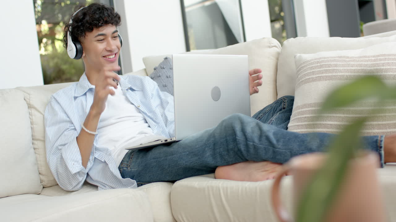 Relaxing on couch, teenage boy using laptop and wearing headphones, enjoying leisure time