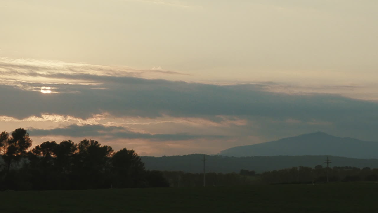 Sunset timelapse of a green land with background mountains in a foggy day. The sun first disappears behind the clouds and its silhouette its revealed