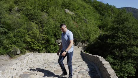 a shot of a tourist looking down the arda river and walking down the devil's bridge on a cobblestone pathway, located in the town of ardino at the foot of rhodope mountains in bulgaria