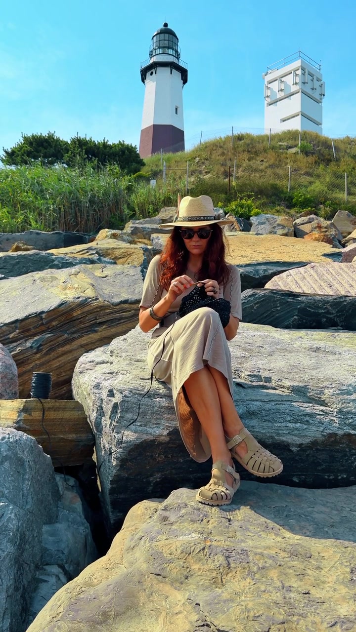 Donna con cappello da sole che lavora all'uncinetto un progetto fatto a mano in riva al mare. Vista frontale di una donna seduta su delle rocce che indossa occhiali da sole e un cappello, concentrata sul lavoro all'uncinetto con filato nero.