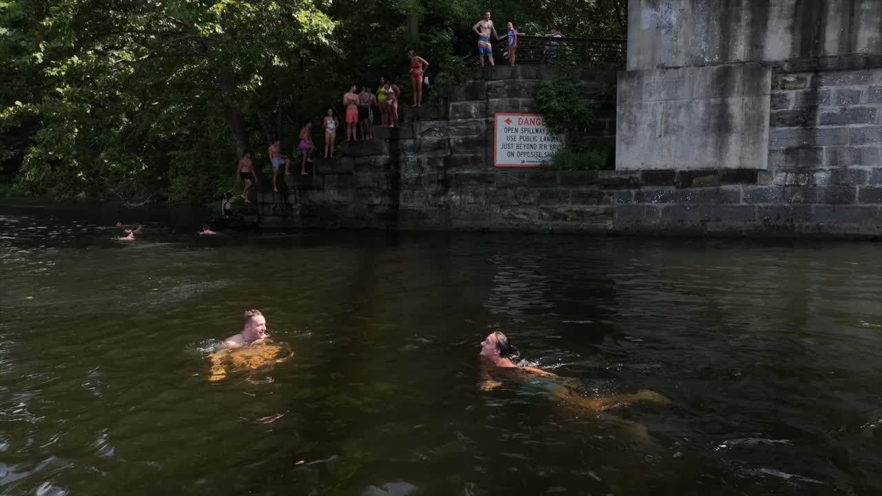 Two friends swimming in James River during sunny day in America. Group of teenager in background Jumping from Bridge. Drone shot.