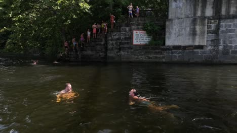 two friends swimming in james river during sunny day in america. group of teenager in background jumping from bridge. drone shot.