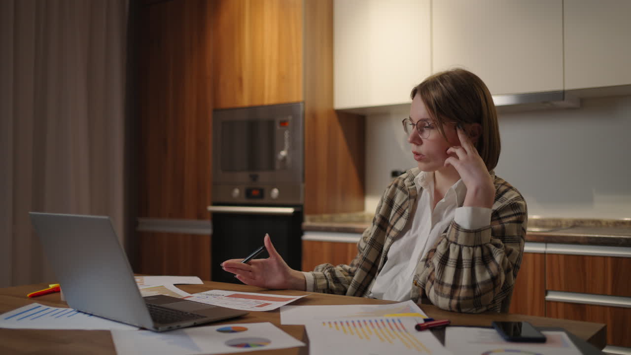 una mujer pensativa y cansada con gafas trabajando en documentos financieros sentada en el lugar de trabajo usando una aplicación de computadora portátil parece concentrada mientras completa una tarea.