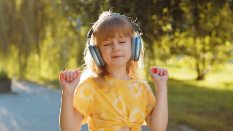 happy child girl in wireless headphones choosing listening music in smartphone dancing outdoors