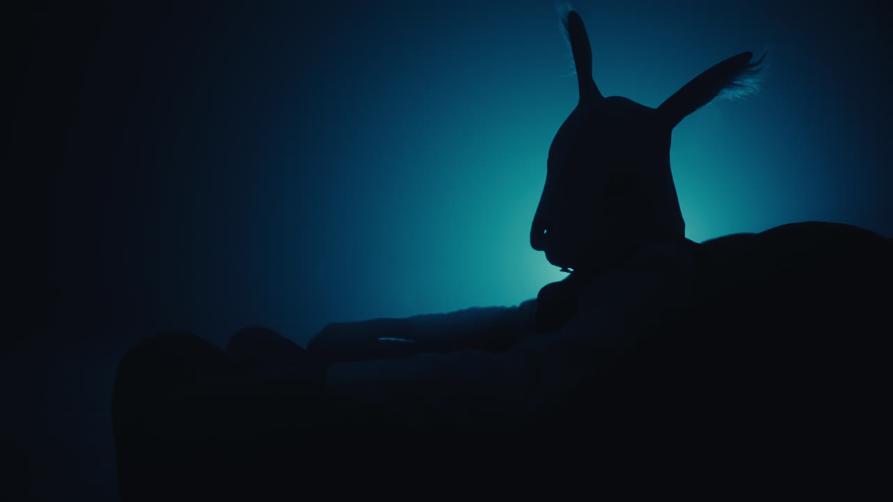 Silhouette of a person wearing a rabbit mask in a dark room lit by blue light