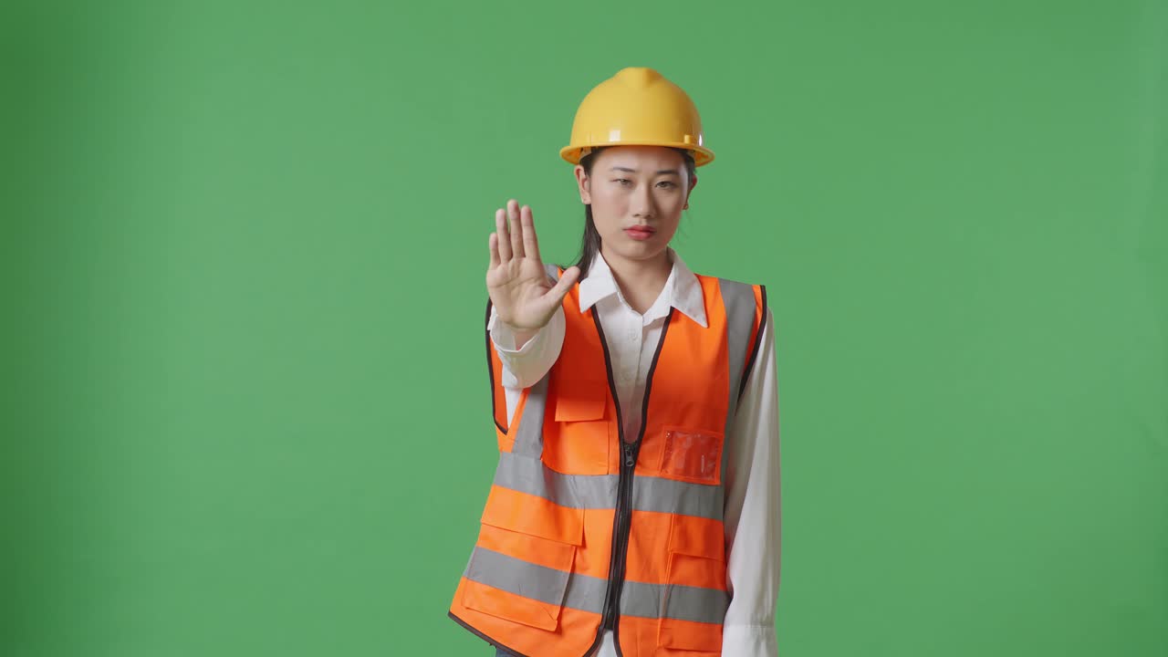 안전 헬을 입은 아시아 여성 엔지니어가 녹색 스크린 배경 스튜디오에 서서 손 표시 없이 불쾌감을 표합니다.