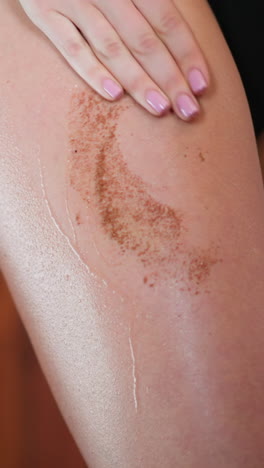 young slim woman spreads brown coffee scrub on gentle skin of thigh with hand making massage in circular motion skincare routine closeup slow motion