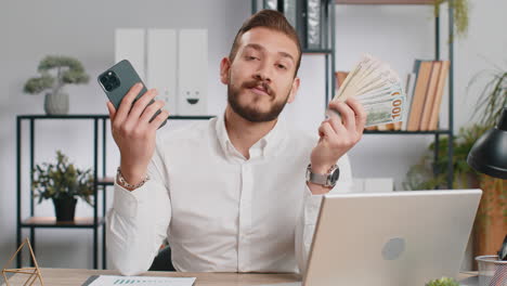 hombre de negocios mostrando teléfono inteligente sinceramente regocijándose ganar, recibiendo dinero dólares billetes en efectivo