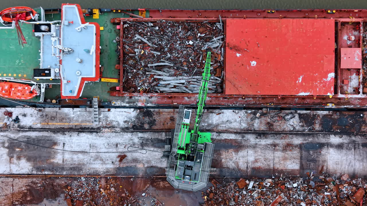 Vista dall'alto di una nave che carica rottami metallici al molo. Prospettiva dall'alto di una gru verde che riempie la stiva di una nave con varie parti metalliche.