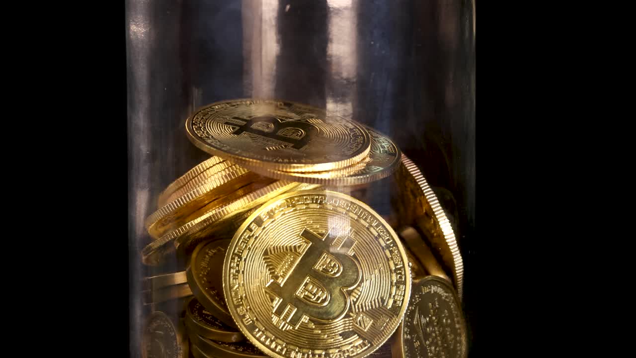 Multiple gold cryptocurrency coins fall into a clear jar, illuminated by warm, focused lighting