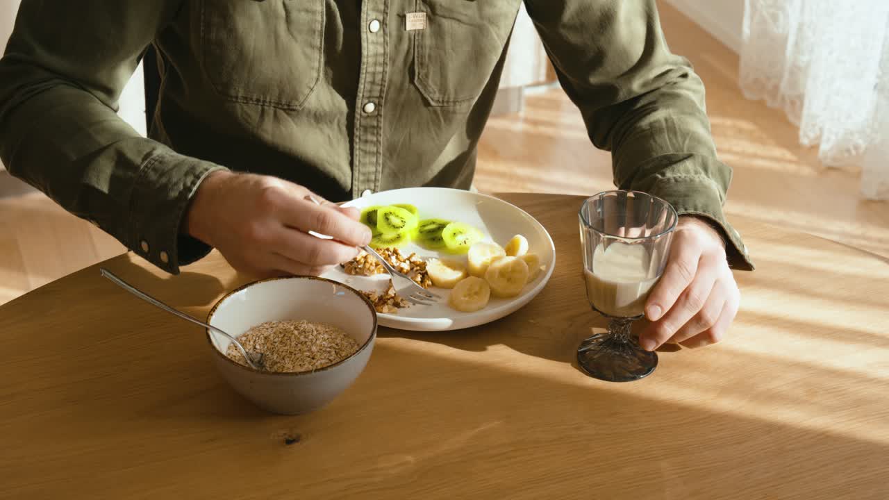 A man sits at the dining table and eats a cereal, bananas and kiwis and drinks a glass of milk. the sun shines on the table Close up