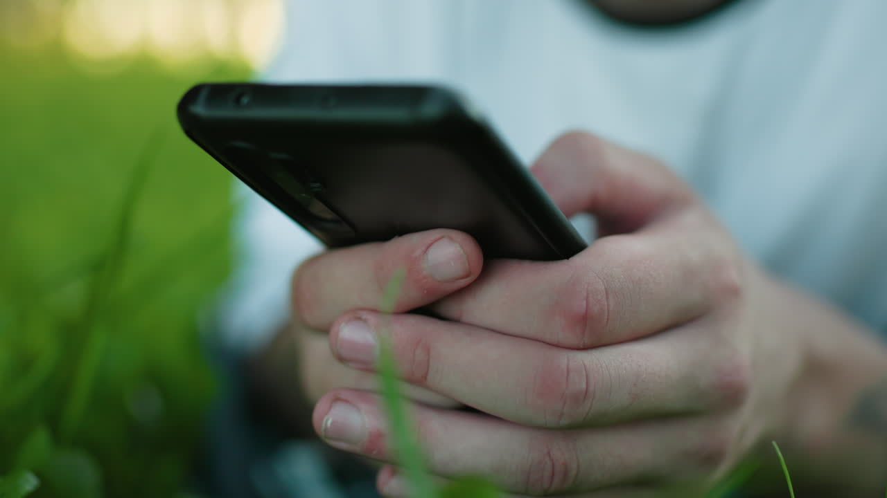 primer plano de un hombre con una camiseta blanca acostado en un campo cubierto de hierba mientras sostiene y opera un teléfono inteligente, centrarse en las manos agarrando el teléfono, hierba verde vibrante en un fondo borroso