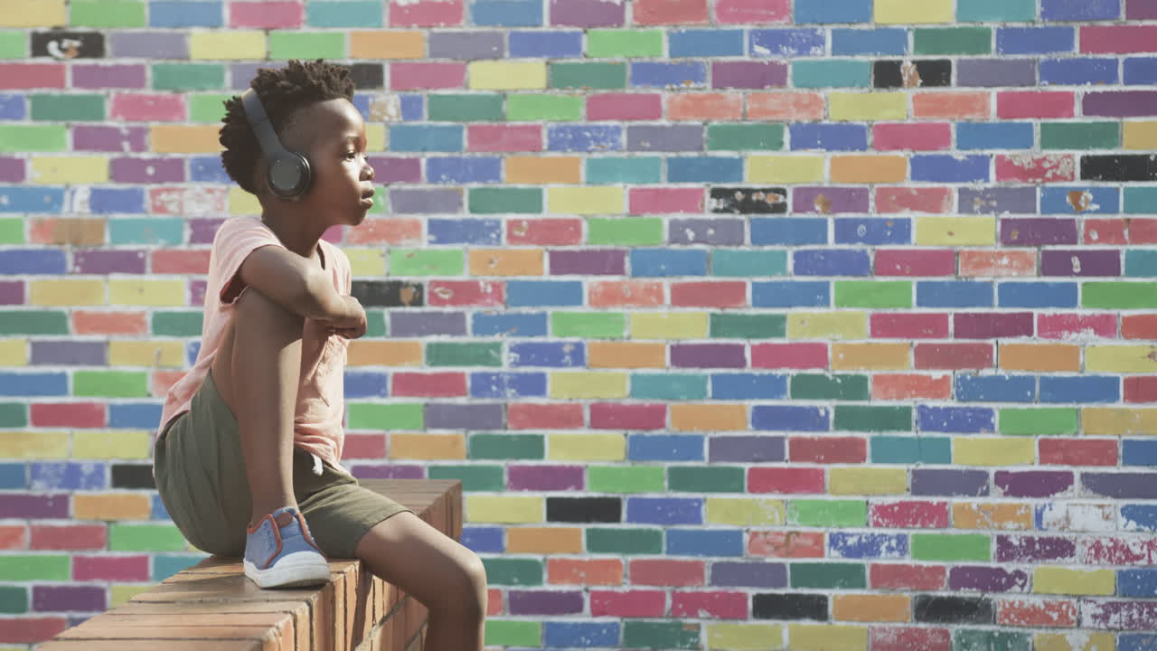 Ragazzo che ascolta musica con le cuffie, seduto su un muretto vicino a un muro di mattoni colorato, con spazio per il testo.