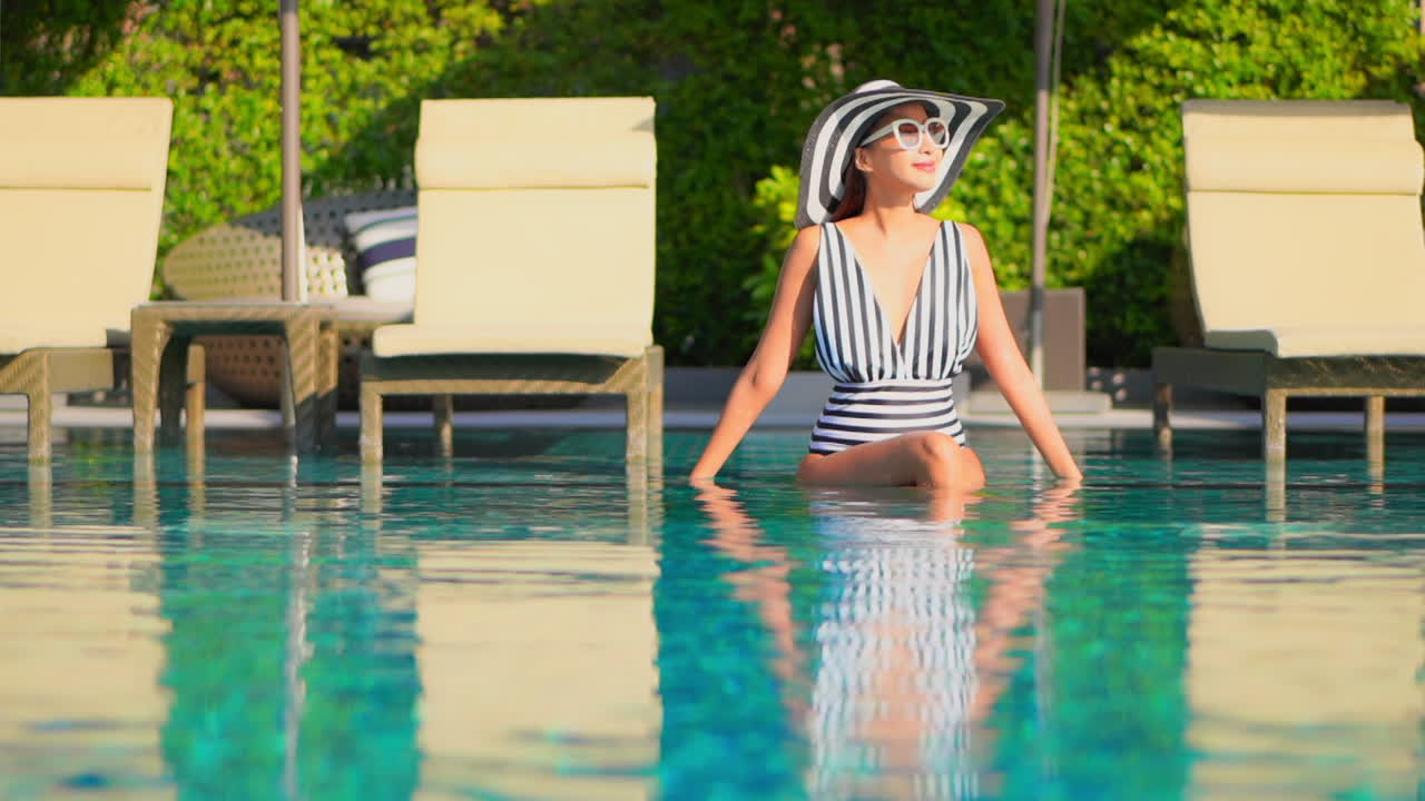 Summer fashion concept, trendy asian woman with black and white hat and swimsuit sitting on swimming pool edge on sunny day, full frame