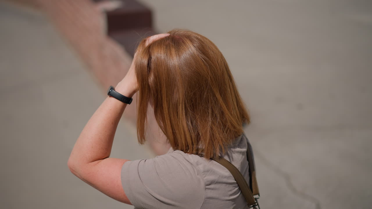 Top view of woman standing beside curb scratching hair with both hands wearing smart watch, showing stress or discomfort, bright daylight highlights grass and pavement