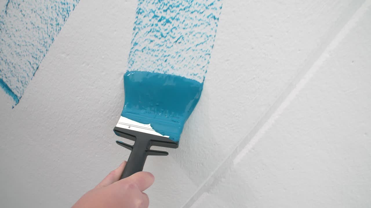 A person's hand paints a white wall with blue paint using a paintbrush