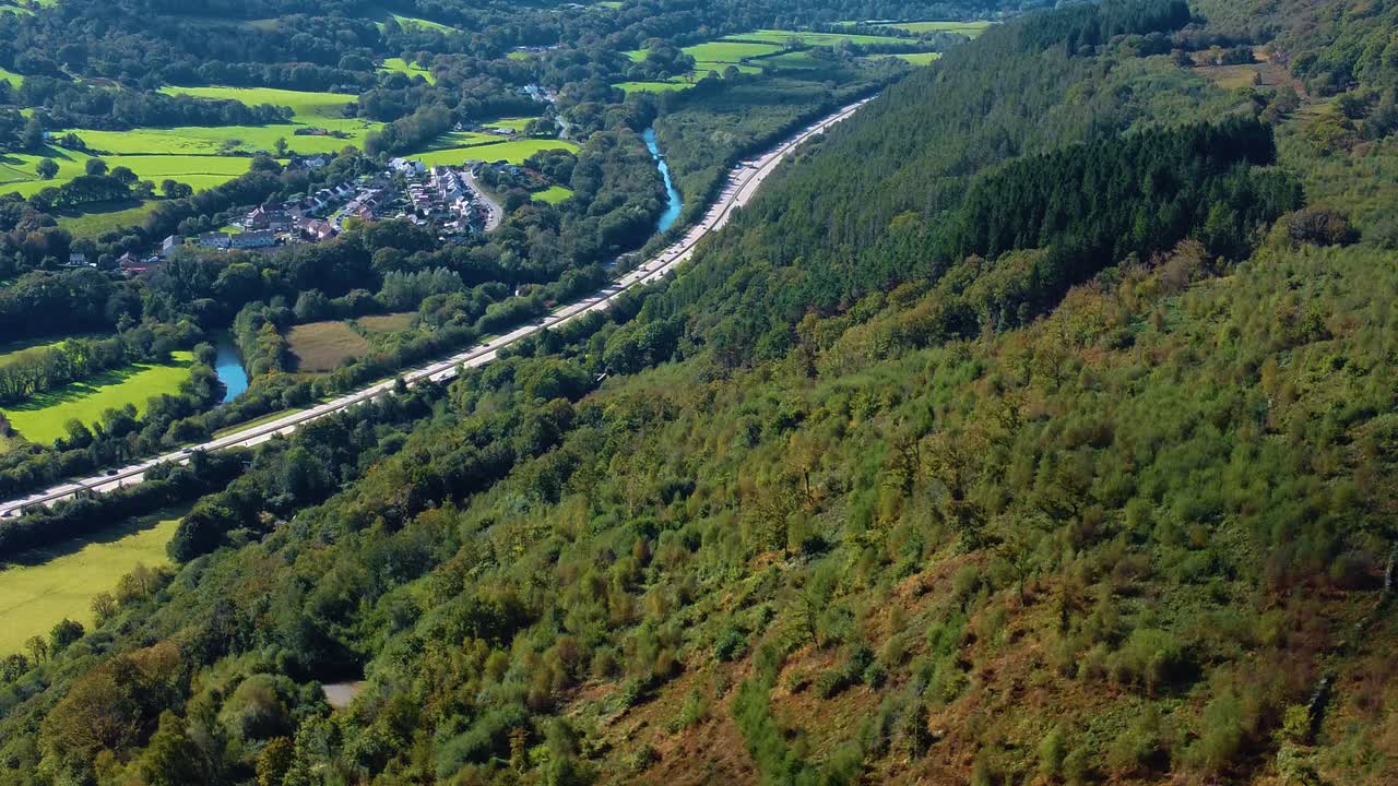 Aerial View Over Forest Covered Hill with Cluster of Residential Houses with Busy Dual Carriageway on A465 Heads of the Valleys Road. Winding River Neath in Background and Open Fields