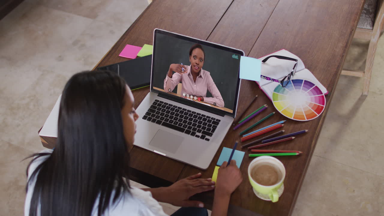 Estudiante caucásica usando una laptop en videollamada con una profesora, tomando apuntes.