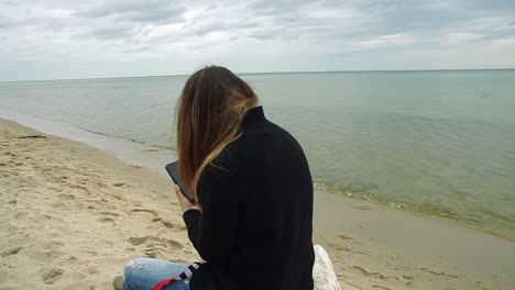The-young-girl-sits-upside-on-a-branch-on-the-seashore-with-her-mobile-phone