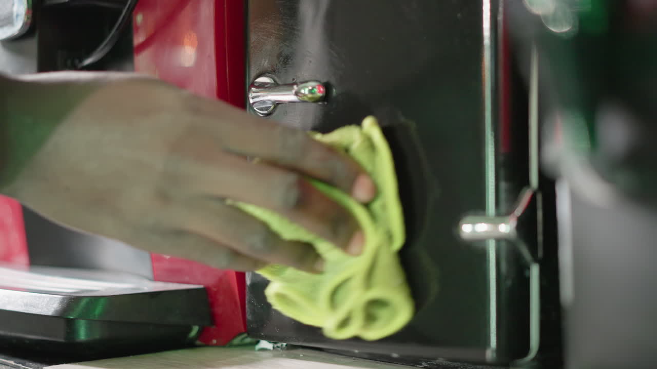 Close-up of hand wiping black surface of red coffee machine with green cloth, focusing on cleaning hard-to-reach areas, ensuring cleanliness and maintenance of kitchen appliance