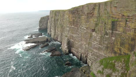 a slow tiliting shot reveals seabirds flying over a turquoise green ocean in front of a dramatic sea cliff rising straight up out of the ocean as waves crash against its base