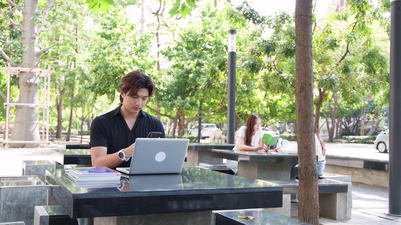 Mahasiswa pria Asia belajar di luar ruangan dengan laptop dan ponsel pintar di bawah cahaya alami yang terang.