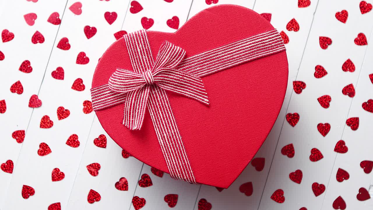 Regalo en caja colocado sobre lentejuelas rojas en forma de corazón en una mesa de madera blanca.
