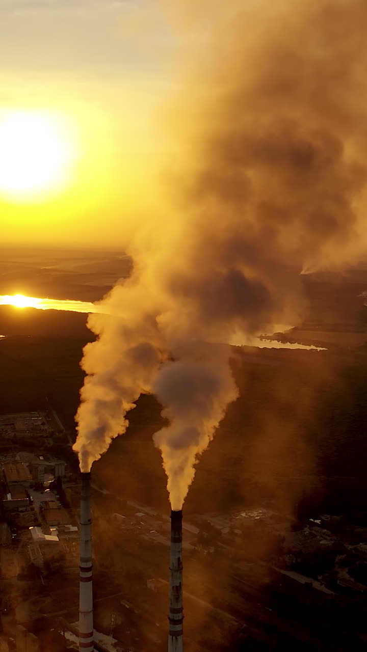 Aerial view on the industrial factory. Power plant near the river on the natural landscape at sunset. Large pipes with smoke in the evening. Vertical video