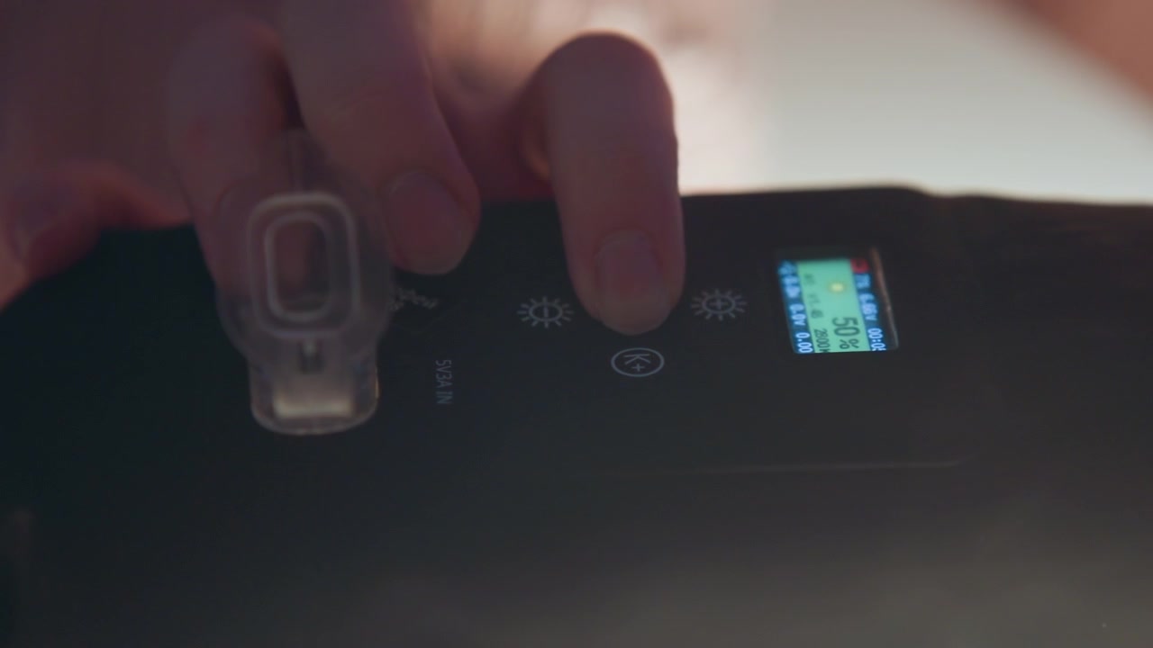 Close-up hand interacts with medical monitoring panel, pressing and rotating controls beside clear tubing connector while illuminated display shows numeric indicators in dim clinical setting