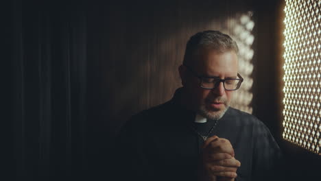 clergyman praying in dimly lit confessional booth
