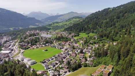 houses and homes with traffic on road during sunny day in laax, switzerland. forest trees in swiss alps. snowy peak at horizon. aerial wide shot