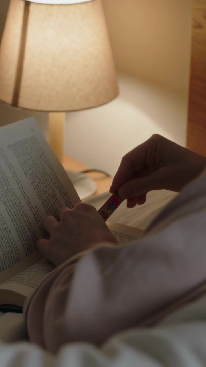 Video verticale: Posizionare un segnalibro sopra un libro, un lettore con un top chiaro che segna la pagina accanto al letto, allungandosi verso la lampada.