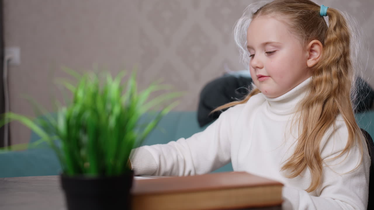 Bambina sorridente con lunghi capelli biondi raccolti in una coda di cavallo, indossa un dolcevita bianco e tiene in mano un quaderno con un disegno mentre è seduta in un ambiente interno vicino a un tavolo con libri e una pianta, attività creativa infantile