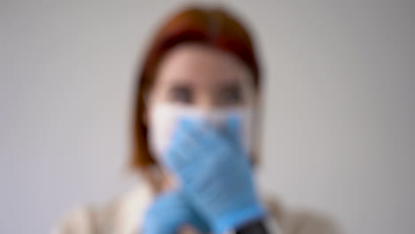 Close-up-of-defocused-woman-putting-on-medical-face-mask-and-looking-at-camera-wearing-gloves.-Copy-space-and-white-background.-Close-up.-Frontal-view