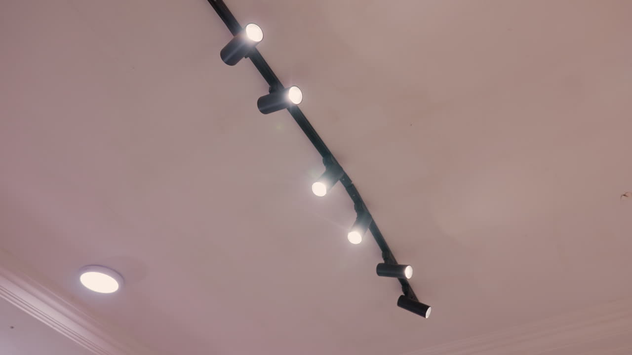 Focos de carril negros en el techo crean un ambiente de galería moderno. Fila de focos ajustables montados en un carril negro, bombillas LED cálidas proyectan haces suaves sobre un techo de yeso y molduras decorativas.