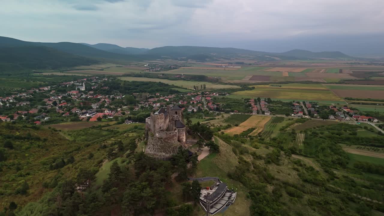 Orbiting aerial view of Boldogkőváralja Castle perched on a hill, surrounded by lush greenery, a quaint village, and scenic Hungarian countryside under a cloudy summer sky