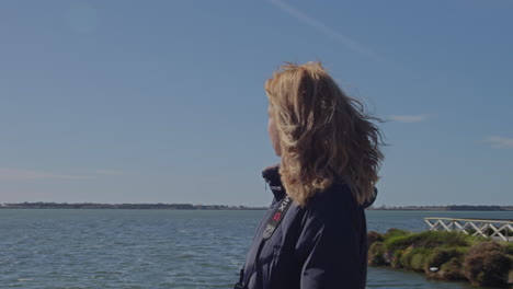 A-girl-walks-along-coast-on-windy-sunny-day,-sea-and-old-lighthouse-behind