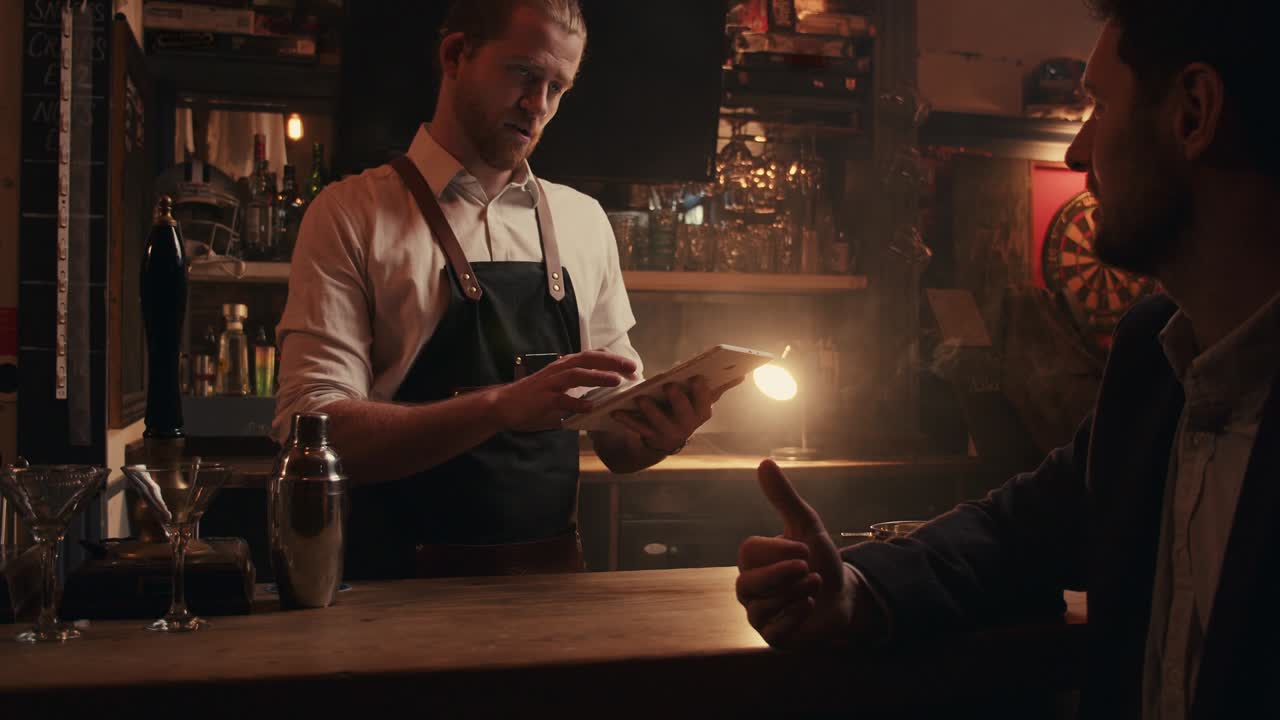 백인 남성 바텐더가 남성 고객과 대화하는 동안 디지털 태블릿에 타이핑하고 있습니다.